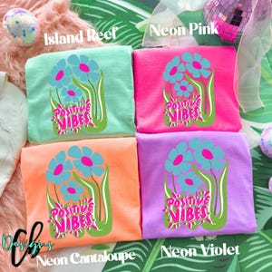 May include: Four colorful t-shirts, each folded and displaying a floral design with the words "Positive Vibes." Colors include Island Reef, Neon Pink, Neon Cantaloupe, and Neon Violet. The shirts have a crew neck.