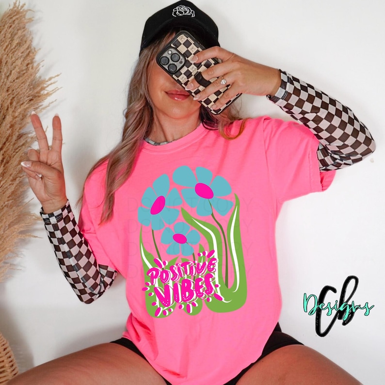 May include: A bright pink t-shirt featuring a floral design with blue and pink flowers and the words "Positive Vibes" in a green and pink font. The person is wearing a black cap and a checkered sleeve.