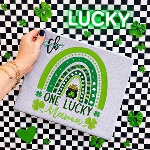 May include: A light gray t-shirt with a green rainbow design, a pot of gold, and the text "ONE LUCKY Mama." The word "LUCKY" is displayed in neon green above the shirt. Green shamrocks and bows decorate the background.