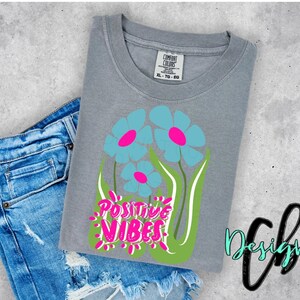 May include: A gray t-shirt with a floral design featuring blue flowers with pink centers and green stems. The words "POSITIVE VIBES" are in pink and white. The shirt is next to a pair of blue denim shorts.