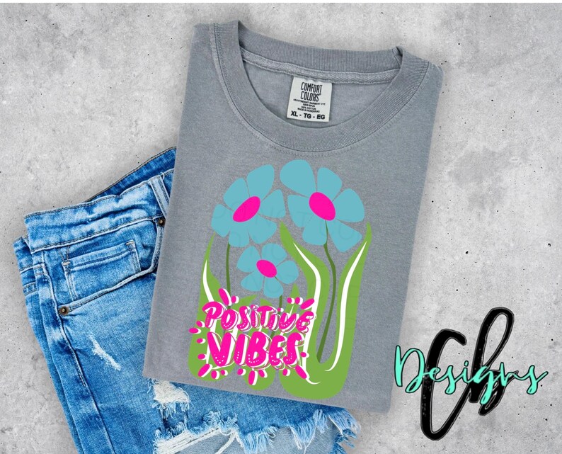 May include: A gray t-shirt with a floral design featuring blue flowers with pink centers and green stems. The words "POSITIVE VIBES" are in pink and white. The shirt is next to a pair of blue denim shorts.