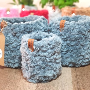 Könnte beinhalten: Drei blaue Häkelkörbe aus weichem, flauschigem Garn. Jeder Korb hat ein kleines braunes Lederetikett, das mit einer silbernen Niete befestigt ist und den Text Made with love trägt. Ein braunes Papieretikett mit dem Logo tellyfinished ist angebracht.