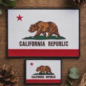 May include: Two rectangular embroidered patches featuring the California state flag design. Each patch displays a white background, a brown grizzly bear walking on a green patch of grass, a red star in the upper left, and the text CALIFORNIA REPUBLIC.