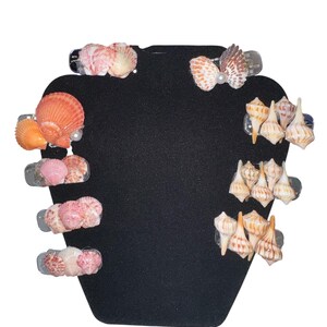 May include: A collection of seashell hair clips in various colors, including pink, orange, and white, displayed on a black background. The clips feature different shell shapes and sizes, some with pearl accents. These accessories are perfect for beach-themed events.
