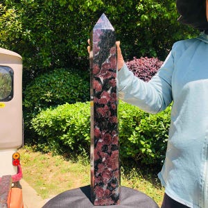 May include: A tall, polished crystal tower featuring a hexagonal prism shape with a pointed top. The stone displays a mottled pattern of deep burgundy red and black mineral inclusions throughout its smooth, reflective surface.