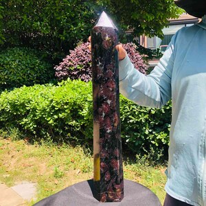 May include: A tall, hexagonal crystal tower featuring a polished surface with a mottled pattern of deep burgundy, black, and white mineral inclusions. The stone has a pointed, faceted top and a solid, flat base, displaying natural variations.
