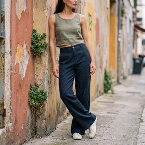 May include: A woman wearing a green tank top and navy blue wide-leg pants. The outfit is paired with white sneakers. The clothing appears to be made of linen. The background is a weathered, colorful wall and a narrow street.