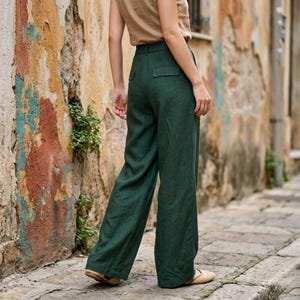May include: A person wearing a tan top and dark green wide-leg pants. The pants have a high waist and a relaxed fit. The background features a weathered, colorful wall and a cobblestone path.