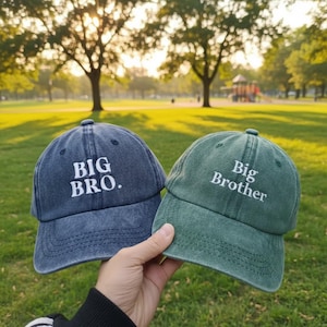 Puede incluir: Dos gorras de béisbol, una azul marino y otra verde, se sostienen sobre un fondo de parque. La gorra azul marino dice "BIG BRO." en texto blanco, mientras que la gorra verde dice "Big Brother". Las gorras están hechas de un material suave y desgastado.