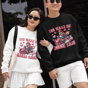 May include: White and black sweatshirts with a cartoon graphic of Mario in a race car, surrounded by hearts. The text "You Make My Heart Race" is printed above and below the graphic in pink. The sweatshirts are worn by two people.