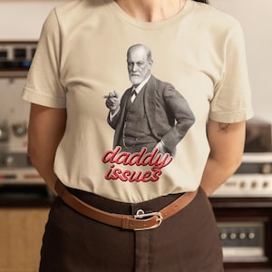 May include: Beige t-shirt featuring a black and white image of Sigmund Freud holding a cigar. The words "daddy issues" are written in red script below the image. The person is wearing a brown belt and brown pants.