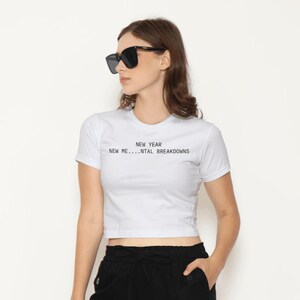 Puede incluir: Top corto blanco con el texto "NEW YEAR NEW ME...NTAL BREAKDOWNS" en negro. La modelo lleva gafas de sol negras y pantalones negros. La camiseta es una prenda casual para el día a día.