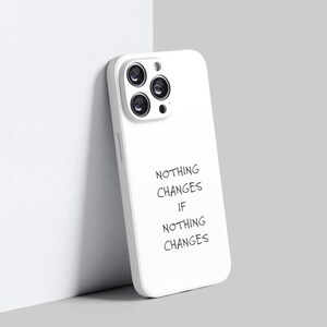 Puede incluir: Una funda blanca para teléfono con un recorte para una cámara de triple lente. La funda se apoya contra una pared blanca y presenta el texto "NOTHING CHANGES IF NOTHING CHANGES" en una fuente manuscrita.