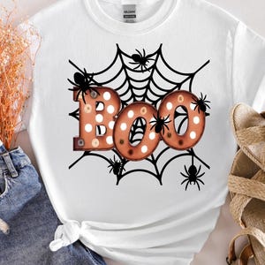 May include: A white short-sleeve t-shirt featuring a graphic of a black spider web with several black spiders and the word BOO in large, orange, polka-dotted letters that resemble vintage marquee lights with visible light bulbs.