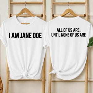 Op de afbeelding: Een wit T-shirt met korte mouwen en zwarte tekst. De voorkant toont de zin I AM JANE DOE in een vetgedrukt schreefloos lettertype. De achterkant toont de zin ALL OF US ARE, UNTIL NONE OF US ARE in hetzelfde vetgedrukte zwarte lettertype.