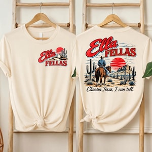 Puede incluir: Una camiseta color crema con un estampado gráfico. El pecho muestra un sombrero de vaquero, un cactus y el texto "Ella FELLAS" en rojo y azul. La espalda muestra un vaquero a caballo, un paisaje desértico, un sol rojo y "Ella FELLAS Choosin Texas, I can tell."
