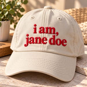 May include: A beige baseball cap featuring the embroidered red text i am, jane doe on the front panel. The cap has a structured crown, a curved brim, and stitched eyelets for ventilation, presenting a clean and simple design.