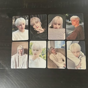 May include: A collection of eight trading cards featuring a person with light blonde hair. The cards display various poses and outfits, including a white sweater, a dark top, and a hooded garment. The cards are rectangular and have a glossy finish.