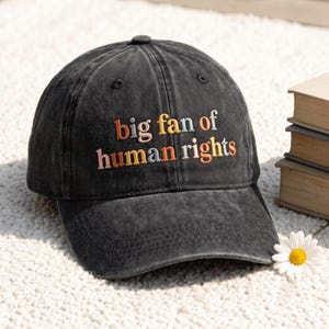 Könnte beinhalten: Eine anthrazitfarbene Baseballkappe mit verwaschener Optik und der gestickten Aufschrift "big fan of human rights" in mehrfarbigen Buchstaben in Orange-, Gelb-, Rosa- und Grautönen auf der Vorderseite der Kappe.