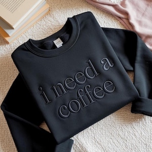 May include: A black long-sleeve crewneck sweatshirt featuring the text "i need a coffee" embroidered in a matching black thread across the chest. The garment has a ribbed collar, cuffs, and hem, with a white brand label visible inside the neckline.