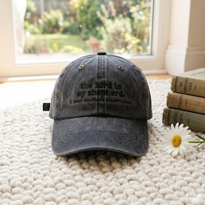 May include: A charcoal gray baseball cap with the embroidered text "the lord is my shepherd. (I need constant supervision.)". The cap has a curved brim and adjustable strap. The cap is sitting on a white rug.