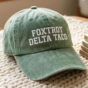 Pu&ograve; includere: Cappellino da baseball verde con la scritta bianca "FOXTROT DELTA TACO". Il cappellino ha una visiera curva e un bottone sulla parte superiore. Realizzato in tessuto morbido e strutturato.
