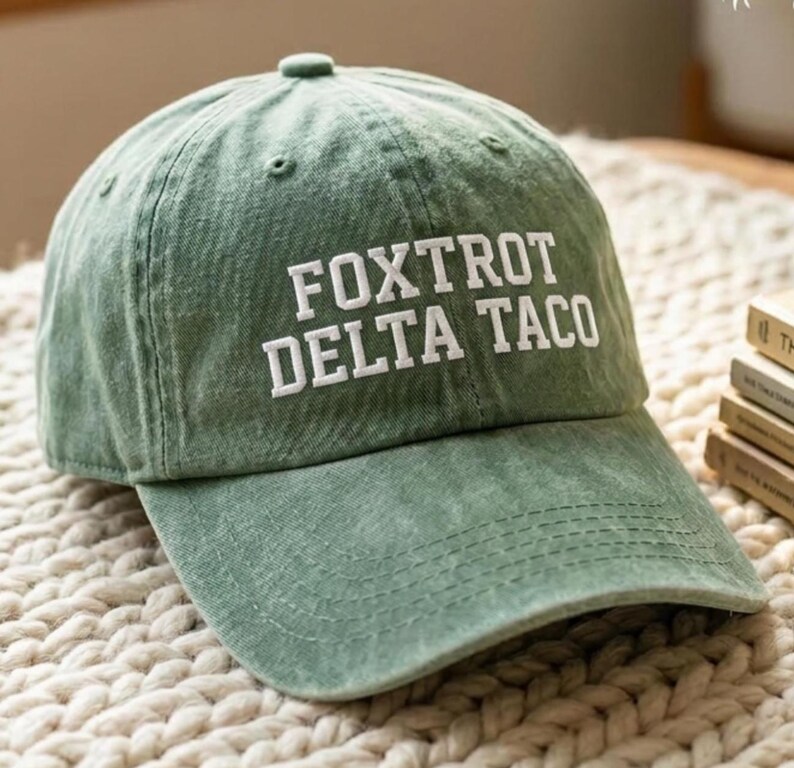 Pu&ograve; includere: Cappellino da baseball verde con la scritta bianca "FOXTROT DELTA TACO". Il cappellino ha una visiera curva e un bottone sulla parte superiore. Realizzato in tessuto morbido e strutturato.