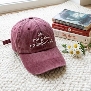 May include: A maroon baseball cap with the text "ok. not good. probably bad." in white. The cap is sitting on a white rug next to a stack of books and a small bouquet of daisies.