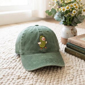May include: A sage green baseball cap featuring an embroidered graphic of a small, light green pickle character with white wings and orange feet centred on the front panel. The cap has a structured crown, a curved brim, and stitched eyelets.