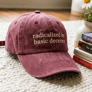 May include: A maroon baseball cap with a distressed fabric finish featuring the embroidered white text "radicalized by basic decency" centered on the front panel. The cap has a curved brim, stitched eyelets, and a button top detail.