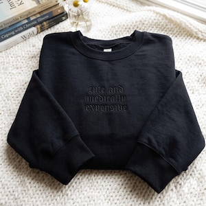Peut inclure: Un sweat-shirt noir à col rond avec le texte brodé "cute and medically expensive" en fil noir assorti. Le vêtement présente des manches longues avec des poignets côtelés et un col côtelé, affichant un design monochrome sur l'ensemble.