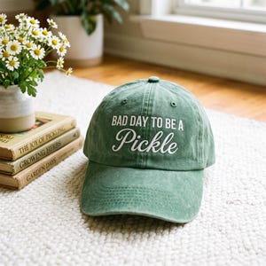 Op de afbeelding: Een saliegroene baseballpet met de geborduurde witte tekst BAD DAY TO BE A Pickle op het voorpaneel. De pet heeft een gestructureerde kroon, een gebogen klep en zichtbare stiksels over het oppervlak van de stof.