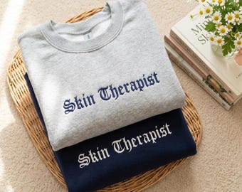 May include: A light gray crewneck sweatshirt with dark blue embroidered text reading Skin Therapist, stacked on a dark navy blue sweatshirt with white embroidered text reading Skin Therapist in a matching gothic-style font.