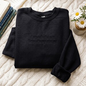 May include: A black crewneck sweatshirt featuring the embroidered text "i speak fluent broadway songs" in a matching black thread. The garment has long sleeves with ribbed cuffs and a ribbed neckline, showcasing a clean and simple design.