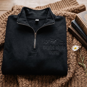 May include: A black quarter-zip sweatshirt featuring an embroidered design on the chest. The embroidery includes a cluster of small flying bats positioned above the text "night shift crew" written in a gothic-style font in matching black thread.