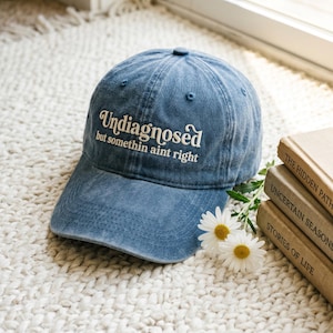 May include: A blue denim baseball cap featuring white embroidered text that reads Undiagnosed but somethin aint right. The cap has a curved brim and a six-panel construction with a button on top, displayed alongside two white daisy flowers.
