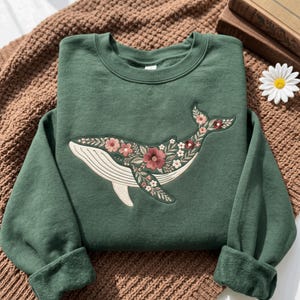 May include: A forest green crewneck sweatshirt featuring a large embroidered graphic of a whale. The whale is filled with a detailed pattern of various flowers, leaves, and vines in shades of pink, red, white, and green, covering its body.