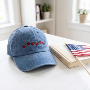 May include: A blue denim baseball cap with an embroidered line of red ladybugs. The cap is sitting on a white surface next to a book and a small American flag.