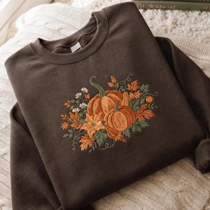 May include: A dark brown crewneck sweatshirt featuring a central embroidered design of two orange pumpkins surrounded by green leaves, small white flowers, and autumn-colored foliage including maple leaves and curling vines on the front chest.