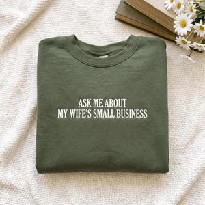 Könnte beinhalten: Ein olivgrüner Rundhals-Pullover mit weißem, mittig auf der Brust gesticktem Text, der lautet: ASK ME ABOUT MY WIFE'S SMALL BUSINESS. Das Kleidungsstück hat lange Ärmel, einen gerippten Halsausschnitt und eine Standardpassform, flach ausgelegt.