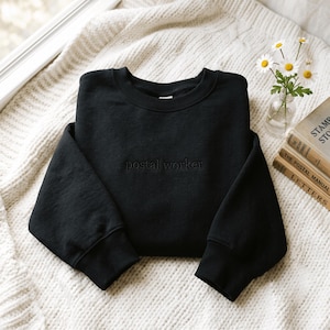 May include: A black crewneck sweatshirt featuring the words postal worker embroidered in a matching black thread across the centre of the chest. The garment has long sleeves with ribbed cuffs and a ribbed neckline, presented in a clean, simple design.