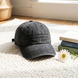 May include: A charcoal gray baseball cap with the embroidered text "I don't rise and shine. I caffeinate and hope." The cap is sitting on a white rug next to a stack of books and a daisy.