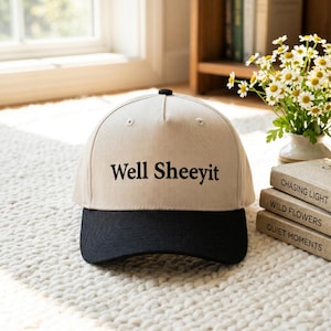 May include: A beige baseball cap with a black brim and a black button on top. The front of the cap features the text "Well Sheeyit" embroidered in black lettering. The cap has a structured crown with ventilation eyelets and a curved brim.