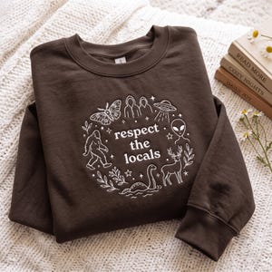 May include: A dark brown long-sleeve sweatshirt featuring a white graphic print of a moth, a UFO, an alien, a deer, a sea creature, and a Bigfoot figure arranged in a circle around the text "respect the locals" with small stars and leaves.