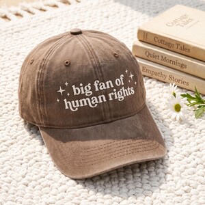 Puede incluir: Una gorra de béisbol marrón con acabado desgastado que presenta el texto blanco "big fan of human rights" impreso en el panel frontal, acompañado de pequeños gráficos de estrellas de cuatro puntas a ambos lados.