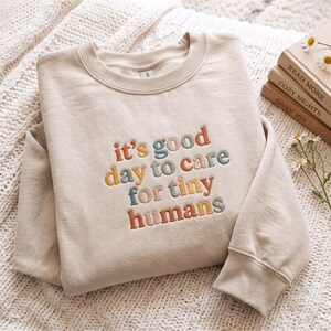 Può includere: Una felpa girocollo color beige con il testo ricamato "it's good day to care for tiny humans" in colori alternati arancione, verde acqua e giallo. Il capo presenta maniche lunghe, un colletto a coste e polsini a coste sulle maniche.