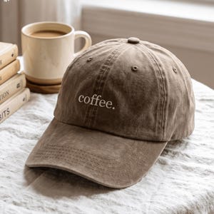 May include: A brown baseball cap with a washed, distressed texture featuring the word coffee. embroidered in white lowercase letters on the front panel. The cap has a curved brim, a button top, and stitched eyelets for ventilation.