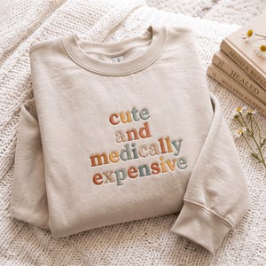 Puede incluir: Una sudadera de cuello redondo en color beige con el texto bordado "cute and medically expensive" en colores alternos de naranja apagado, marrón claro y verde salvia. La prenda tiene mangas largas, cuello acanalado y una textura suave.