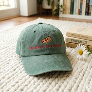 Könnte beinhalten: Eine salbeigrüne Baseballkappe mit gebogenem Schirm. Die Kappe hat einen gestickten Hotdog und den Schriftzug "bad day to be a hot dog" in roter Farbe. Der Hut ist aus einem weichen, strukturierten Stoff gefertigt.