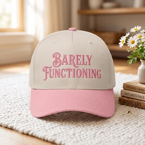 May include: A beige baseball cap with a pink brim and a pink embroidered text on the front that reads "BARELY FUNCTIONING". The cap features a structured crown, a top button, and stitched detailing on the brim, presenting a two-tone color scheme.